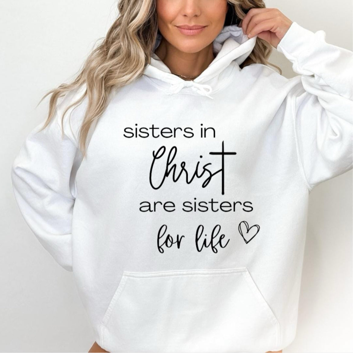 White hoodie that says, “sisters in Christ are sisters for life” with a heart after the saying. There is a cross on the sleeve.