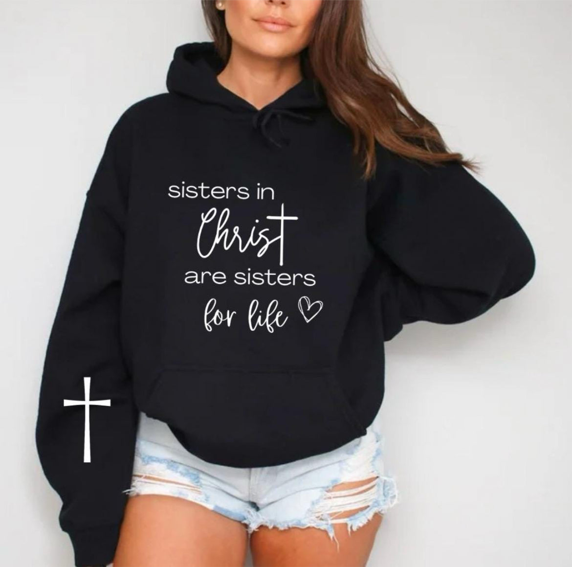 Black hoodie that says, “sisters in Christ are sisters for life” with a heart after the saying. There is a cross on the sleeve.