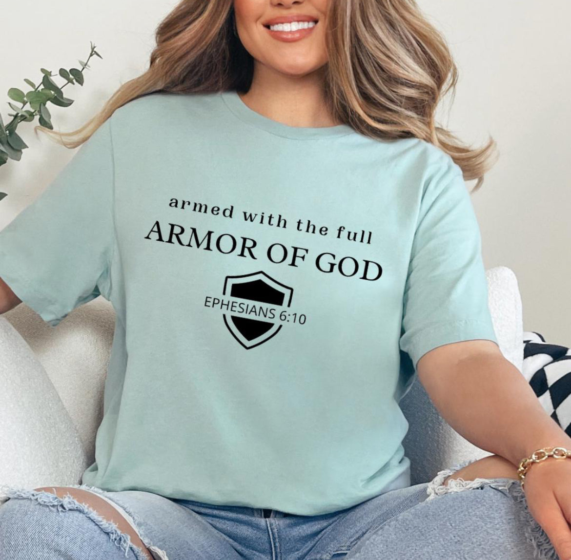 Dusty green crewneck t-shirt that says, “armed with the full armor of God.” There is a shield with “Ephesians 6 10” running through the middle.