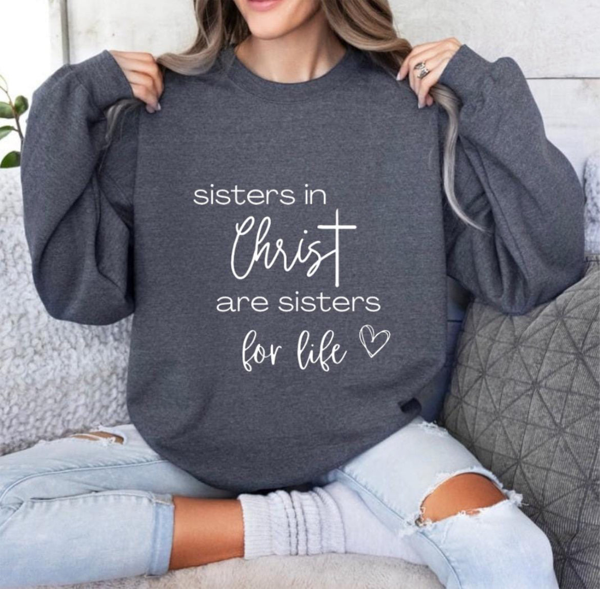 Dark grey crewneck sweatshirt that says, “sisters in Christ are sisters for life” with a heart after the saying. There is a cross on the sleeve.