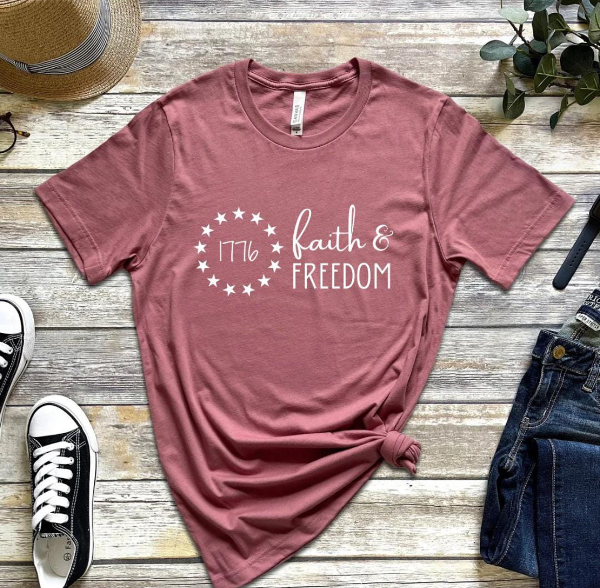 Mauve crewneck t-shirt that says, “Faith and freedom.” There is a Betsy Ross flag stars symbol with “1776” in the middle.