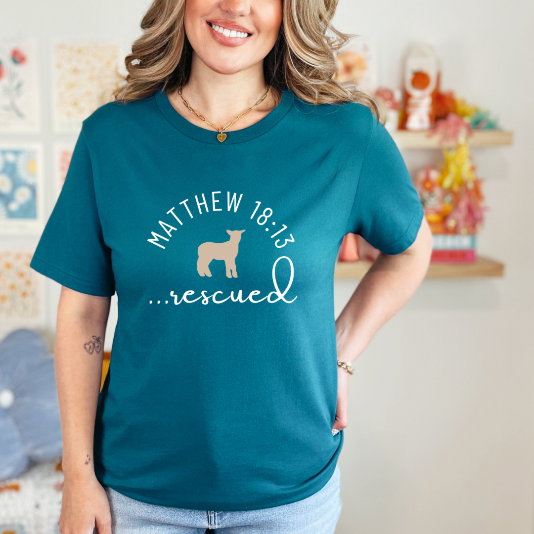 Deep teal crewneck T-Shirt that says, "He left the 99 to rescue me" with a large tan cross. On the front it says, "Matthew 18:13 rescued" with a tan sheep.