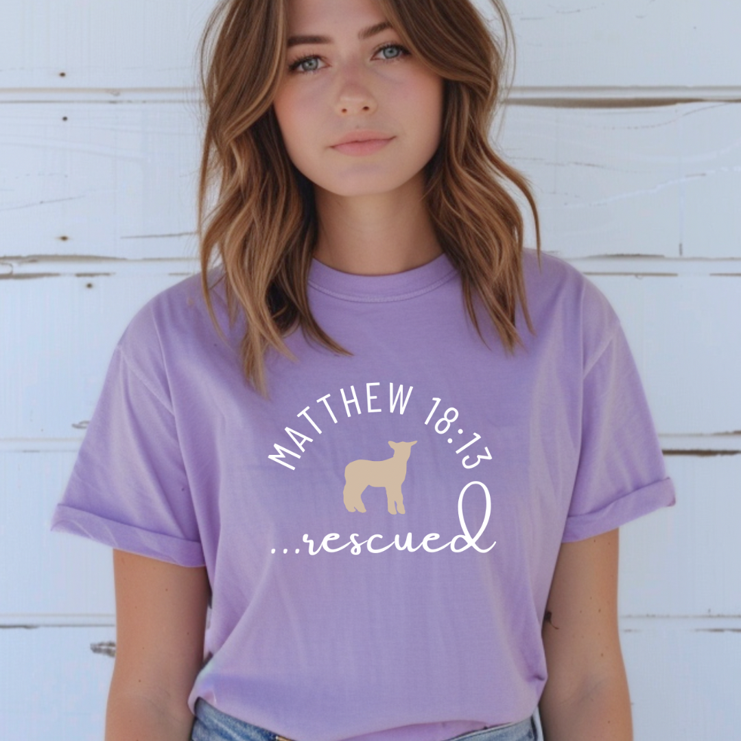 lavender crewneck T-Shirt that says, "He left the 99 to rescue me" with a large tan cross. On the front it says, "Matthew 18:13 rescued" with a tan sheep.