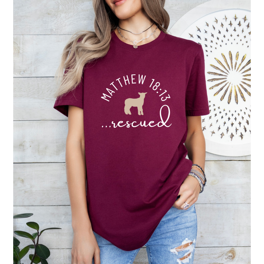 Maroon crewneck T-Shirt that says, "He left the 99 to rescue me" with a large tan cross. On the front it says, "Matthew 18:13 rescued" with a tan sheep.