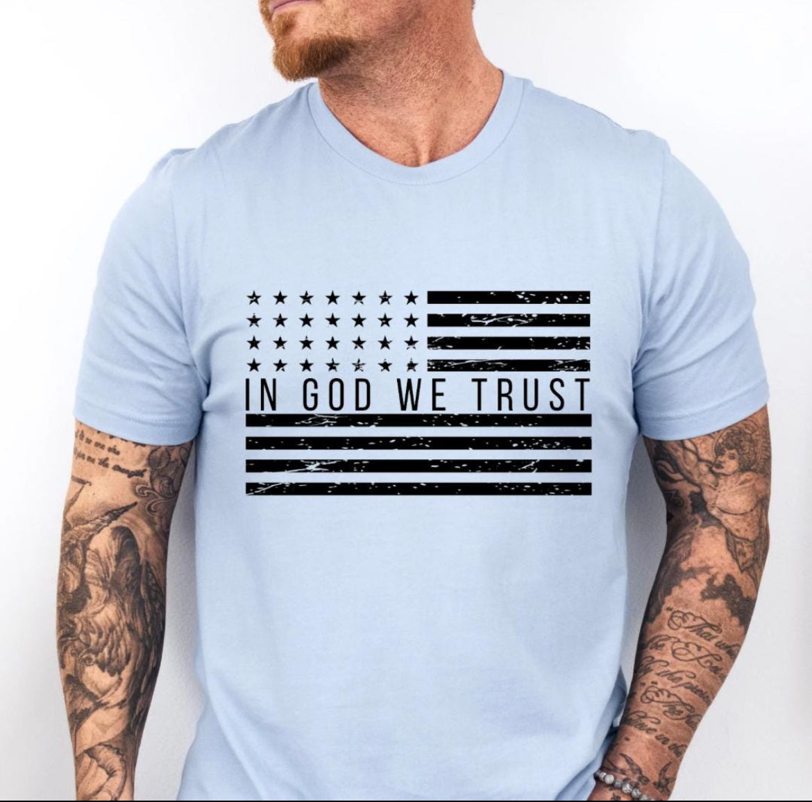 Baby blue crewneck t-shirt that has a distressed American flag. In the middle of the flag it says, “In God we trust.”