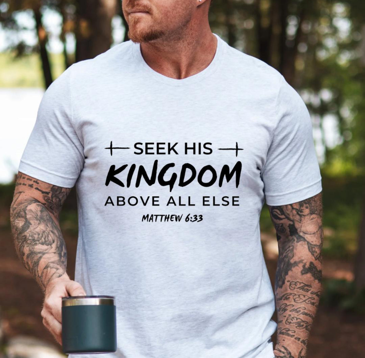 Ash white Crewneck t-shirt that says, “seek his kingdom above all else” in large letters. Underneath in smaller letters it says, “Matthew 6 33.” There are small crosses as embellishments. 