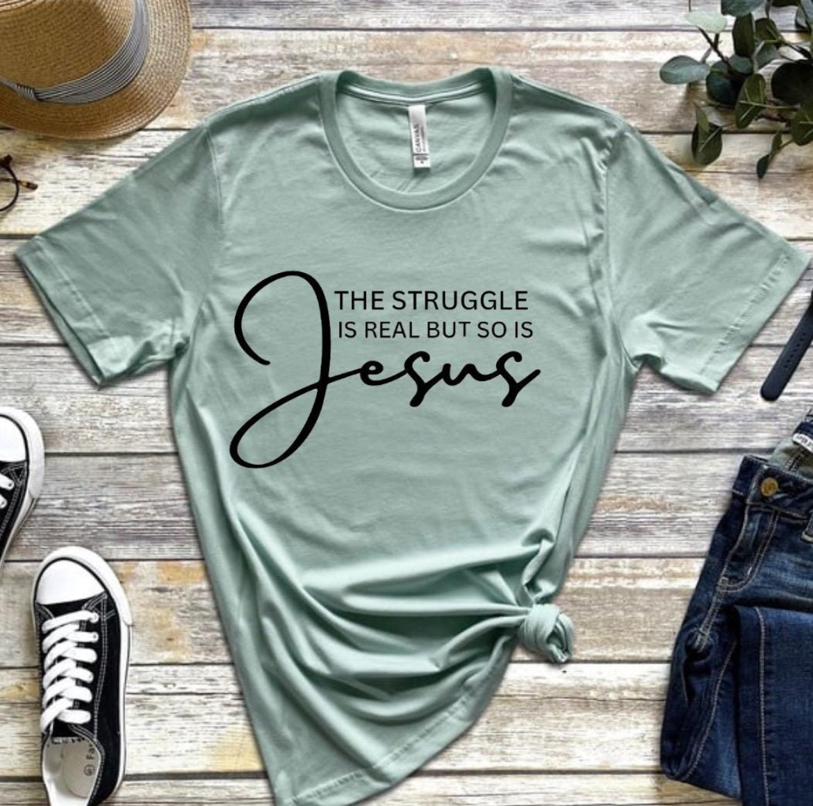 Dust green crewneck t-shirt that says, “the struggle is real but so is Jesus”.