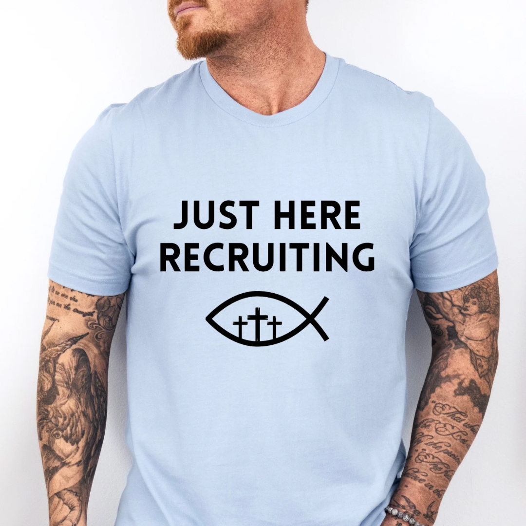 Baby blue crewneck t-shirt that says, “just here recruiting” with the ichthus fish symbol. In the middle of the fish are three crosses.