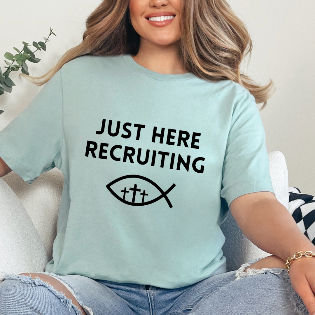 Dusty green crewneck t-shirt that says, “just here recruiting” with the ichthus fish symbol. In the middle of the fish are three crosses.