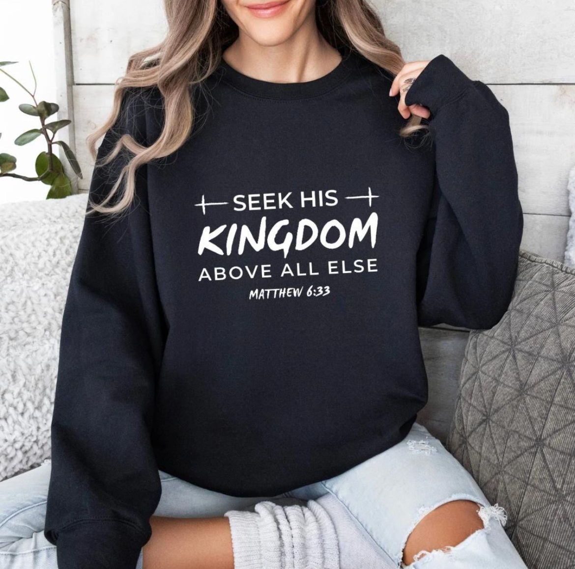 Black crewneck sweatshirt that says, "Seek his Kingdom above all else." Underneath in smaller letters it says, "Matthew 6 33." There are two crosses as embellishments.