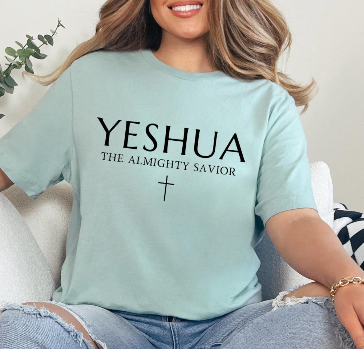 Dusty green Crewneck T-shirt that says, “Yeshua” in large letters. Underneath in smaller letters it says, “the almighty savior” and under that is a small cross. 