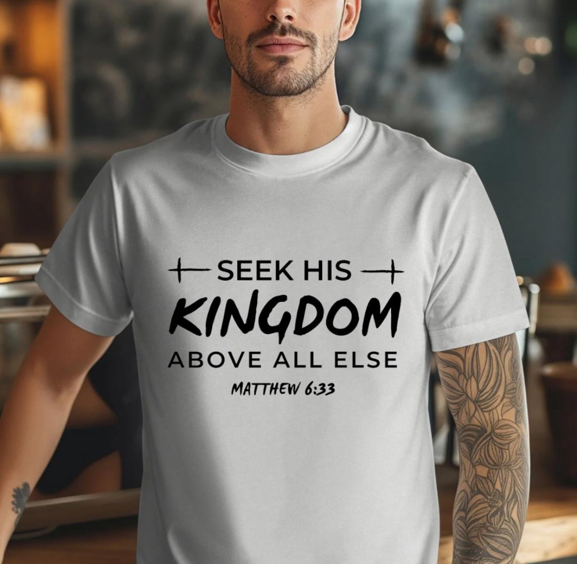 Grey Crewneck t-shirt that says, “seek his kingdom above all else” in large letters. Underneath in smaller letters it says, “Matthew 6 33.” There are small crosses as embellishments. 