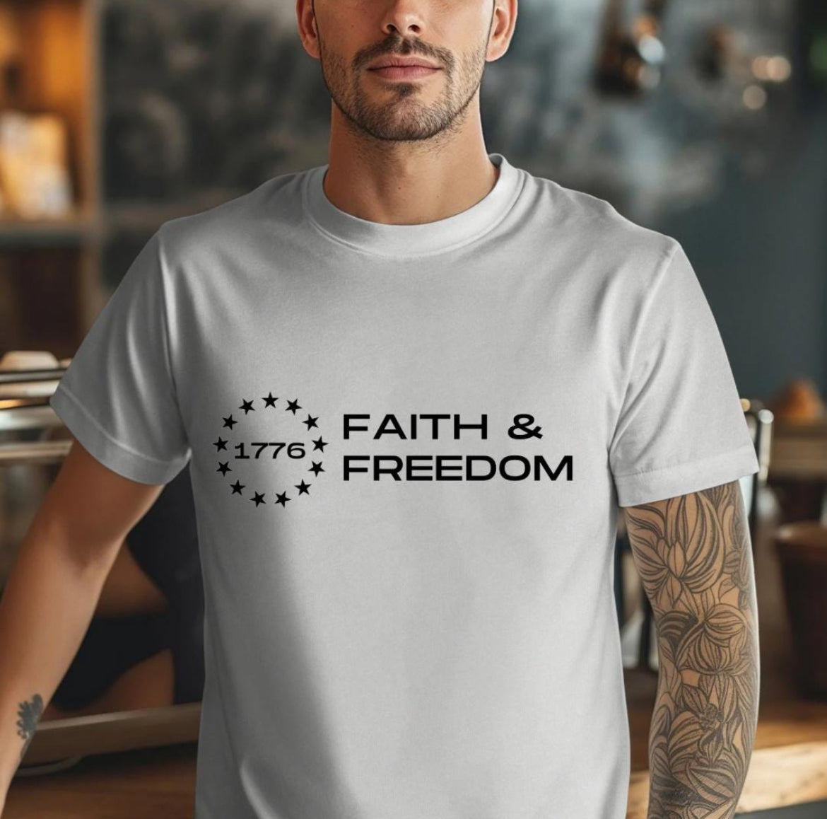 Gray crewneck t-shirt that says, “Faith and freedom” with a Betsy Ross flag star symbol and in the middle it says, “1776”.