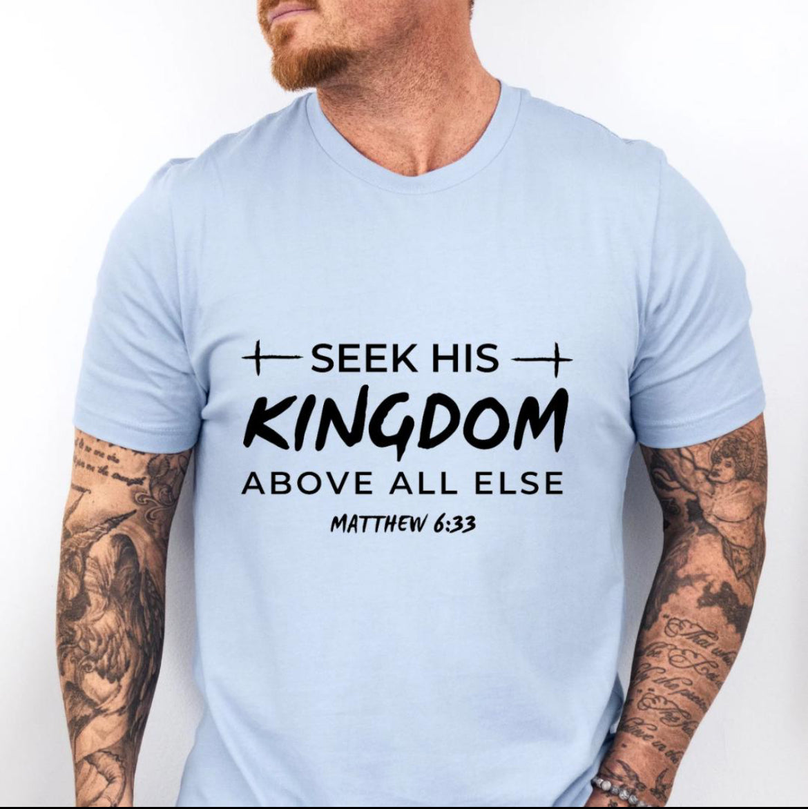 Baby blue Crewneck t-shirt that says, “seek his kingdom above all else” in large letters. Underneath in smaller letters it says, “Matthew 6 33.” There are small crosses as embellishments. 