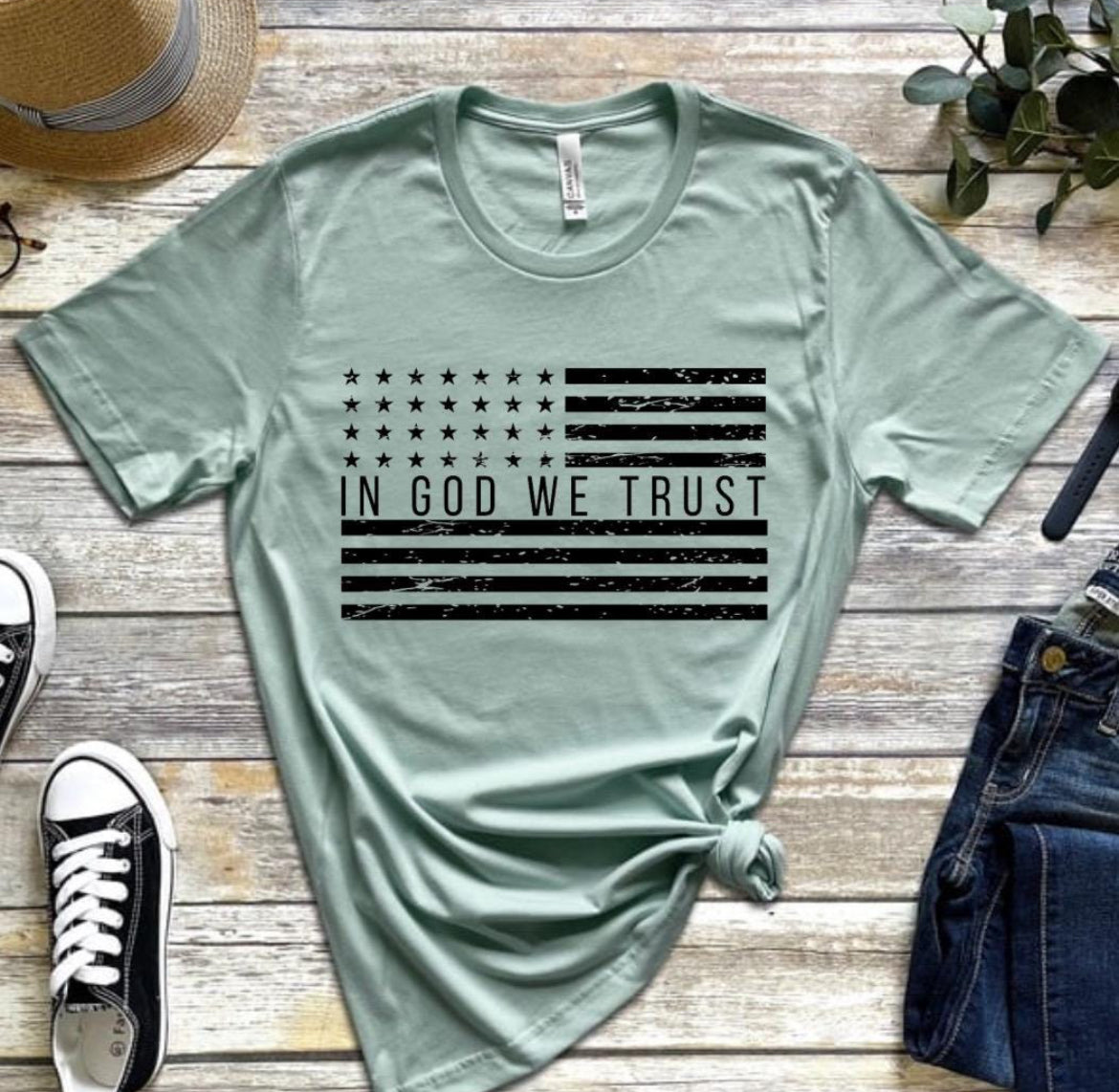 Dust green crewneck t-shirt that has a distressed American flag. In the middle of the flag it says, “In God we trust.”