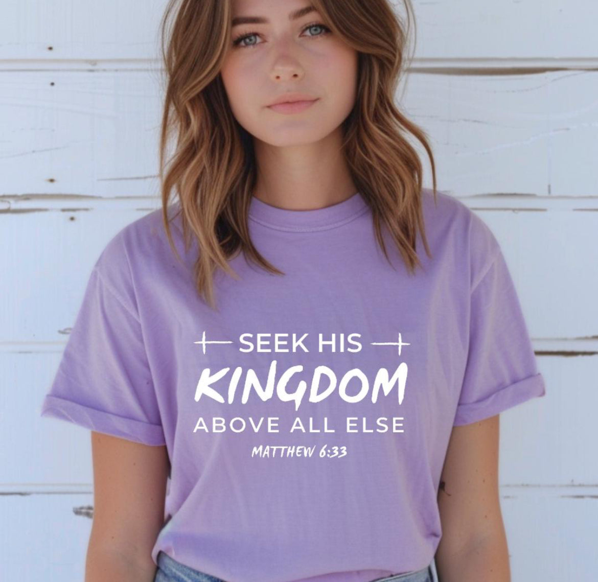 Lavender Crewneck t-shirt that says, “seek his kingdom above all else” in large letters. Underneath in smaller letters it says, “Matthew 6 33.” There are small crosses as embellishments. 