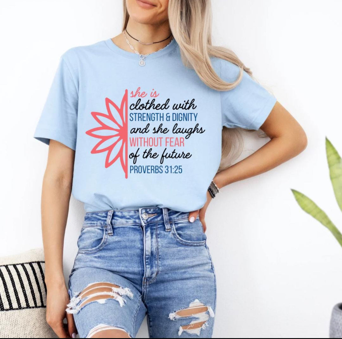 Baby blue crewneck t-shirt that says, “she is clothed with strength and dignity and she laughs without fear of the future. Proverbs 31:25.” There is a large flower. The words are peach, navy and black. 