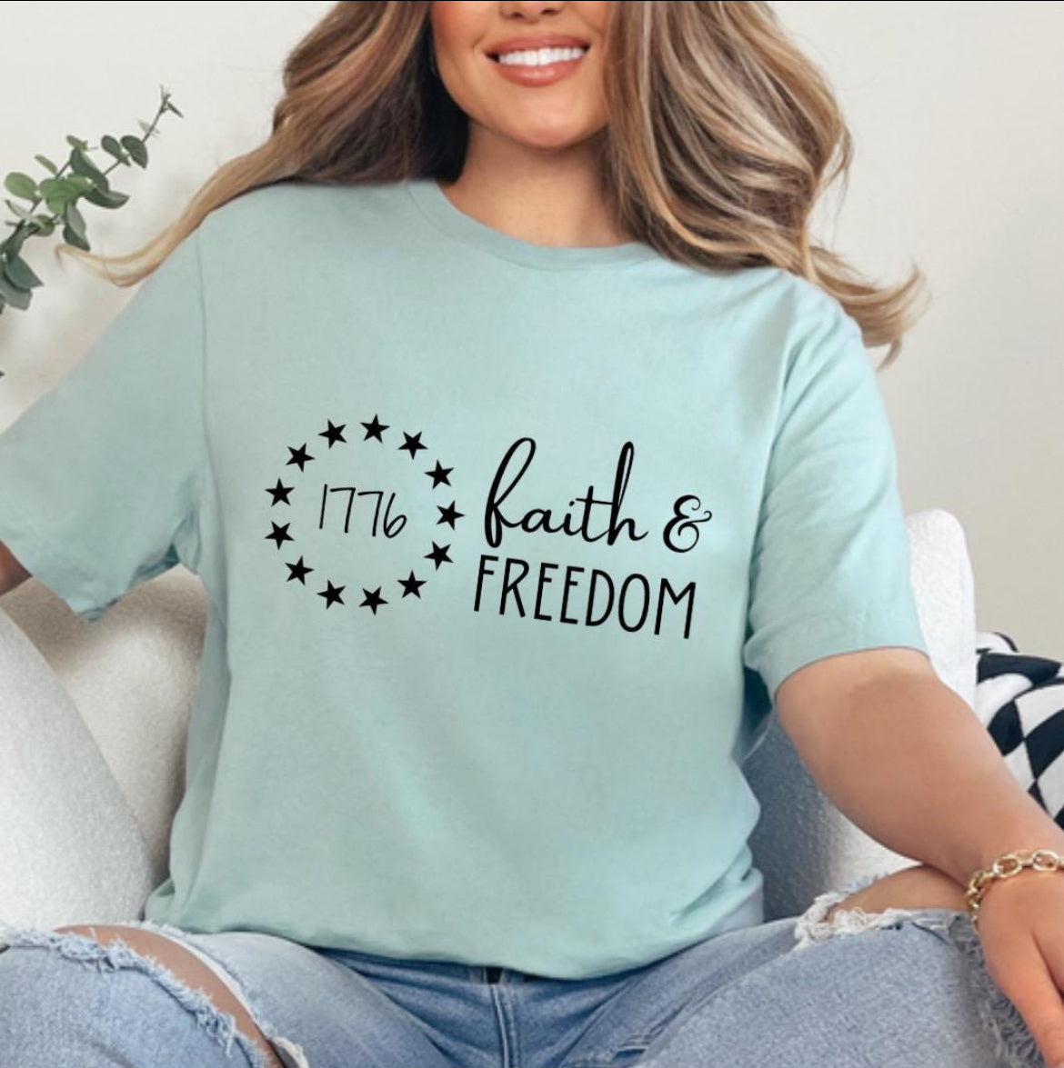 Dusty green crewneck t-shirt that says, “Faith and freedom.” There is a Betsy Ross flag stars symbol with “1776” in the middle. 