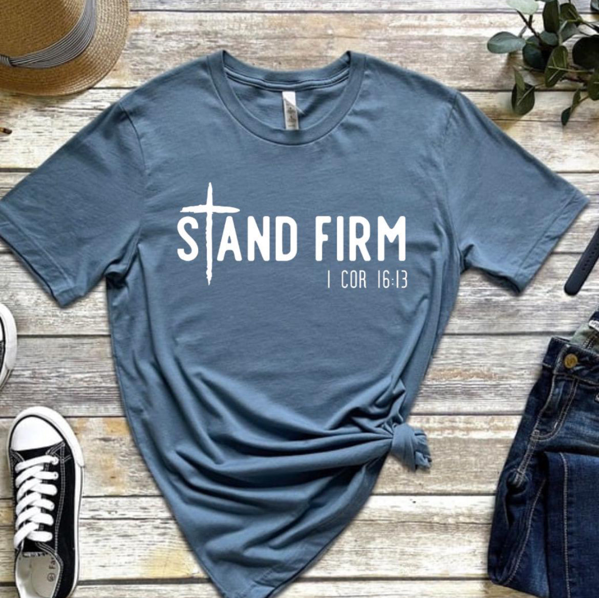 Steel blue Crewneck shirt that says, “Stand Firm” in large letters. The “t” in stand is a cross. Underneath in smaller letters it says, “1 Corinthians 16:13”.