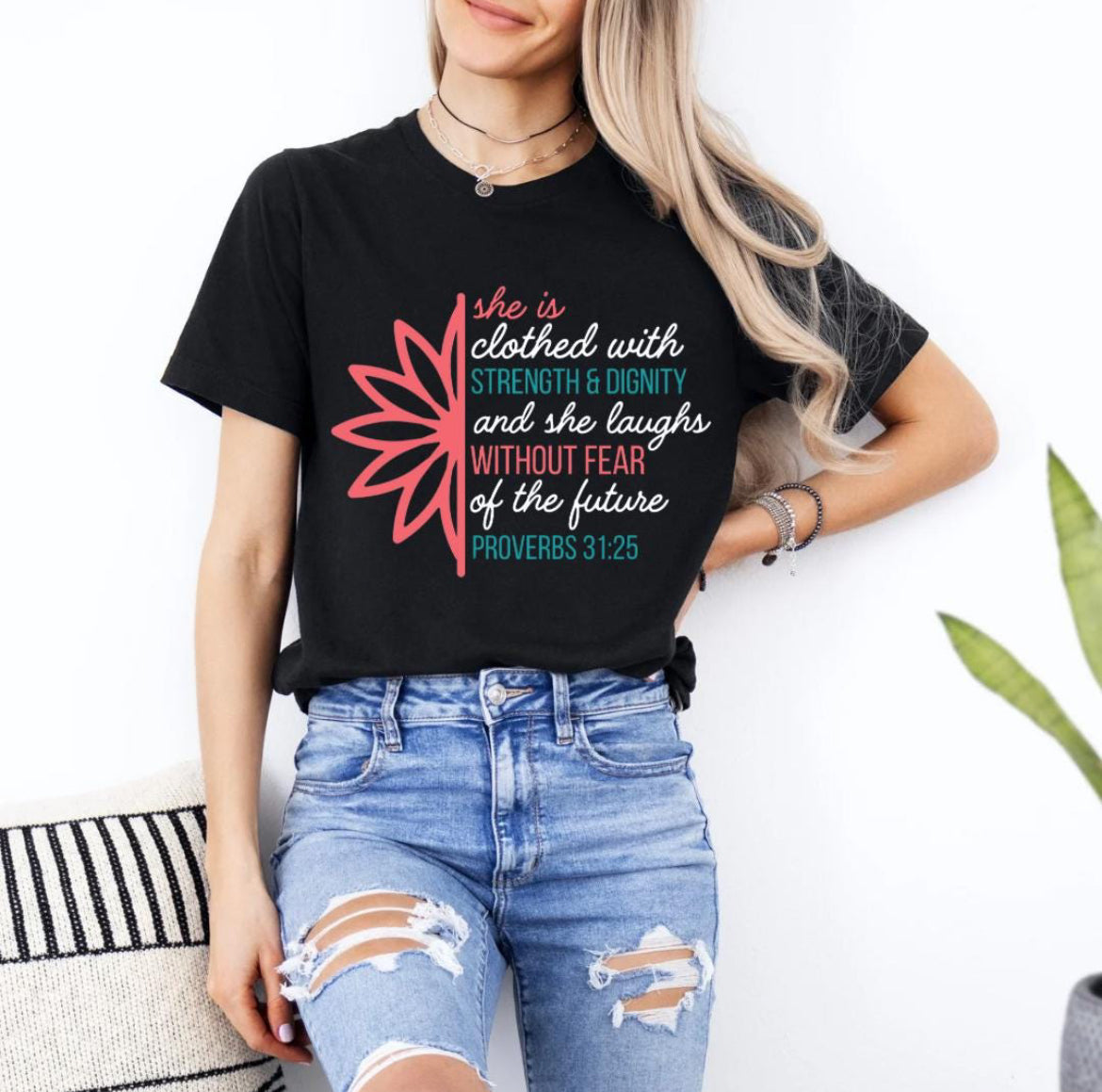 Black crewneck t-shirt that says, “she is clothed with strength and dignity and she laughs without fear of the future. Proverbs 31:25.” There is a large flower. The words are peach, teal and white.