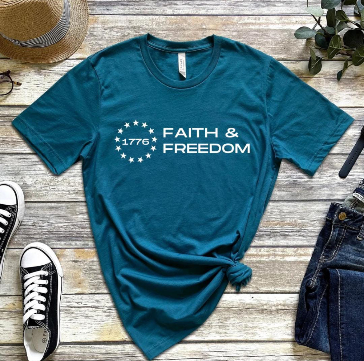 Deep teal crewneck t-shirt that says, “Faith and freedom” with a Betsy Ross flag star symbol and in the middle it says, “1776”.