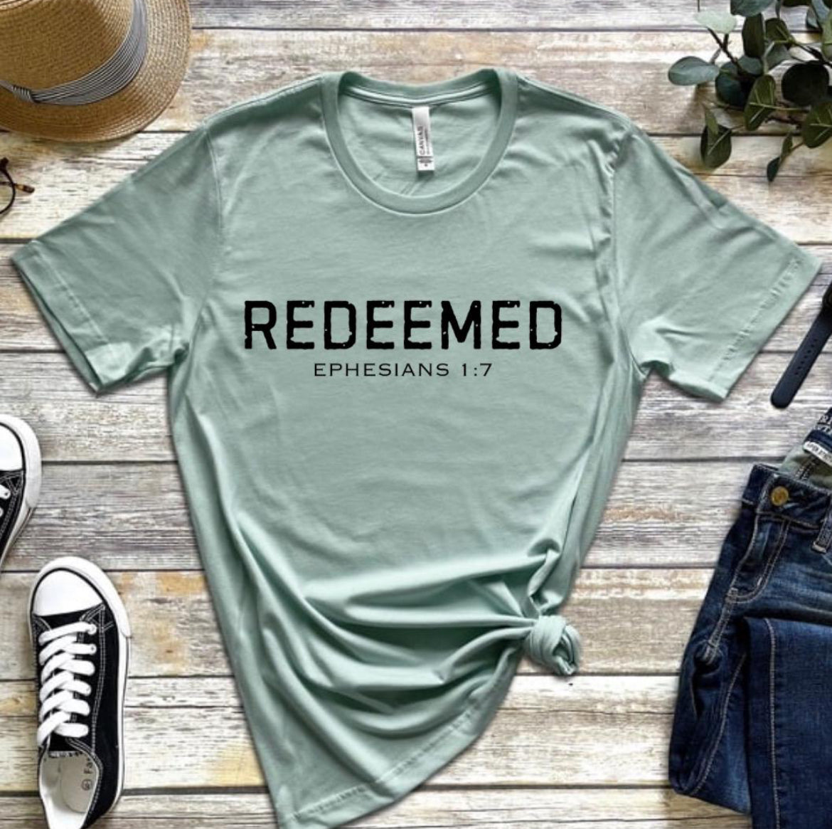 Dusty green Crewneck t-shirt that says, “redeemed” in large distressed letters. Underneath in smaller letters it says, “Ephesians 1 7.”