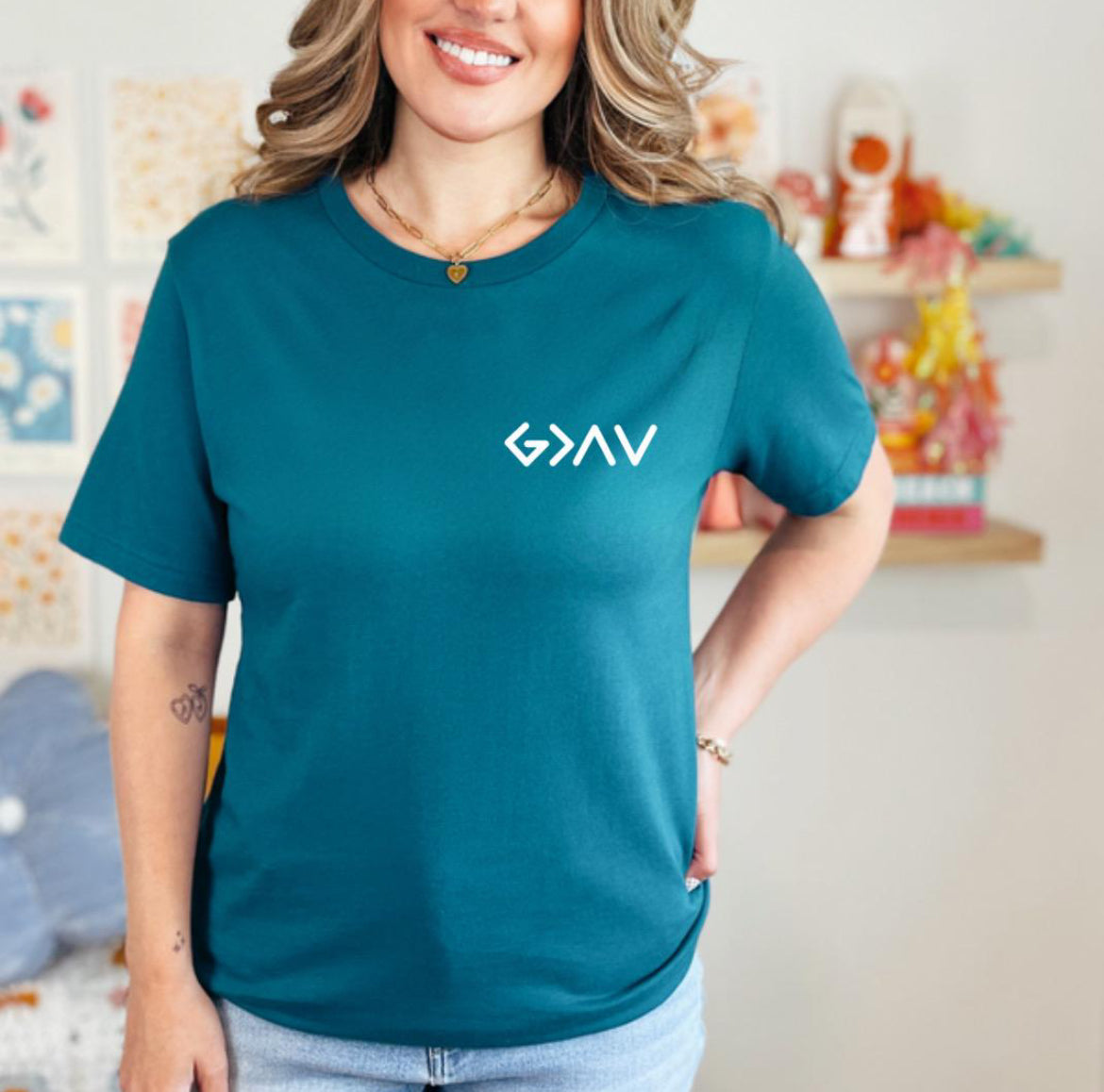 Deep teal crewneck t-shirt with a cross on the back that has the symbol “God is greater than the highs and lows” as the bottom part of the cross.  The front has the same symbol in the upper left-hand corner.