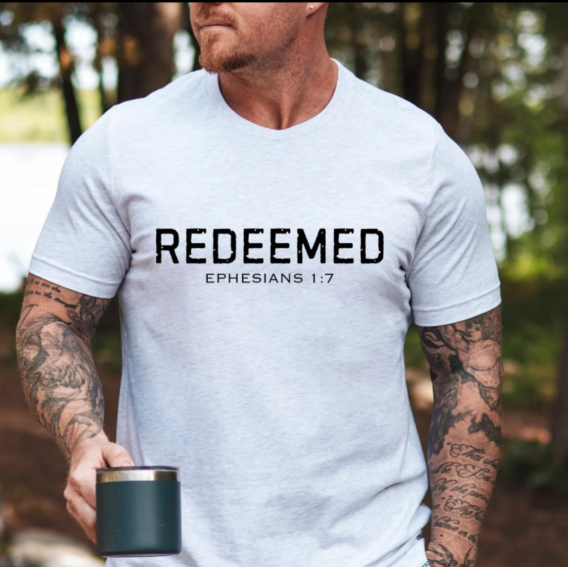 Ash white Crewneck t-shirt that says, “redeemed” in large distressed letters. Underneath in smaller letters it says, “Ephesians 1 7.”