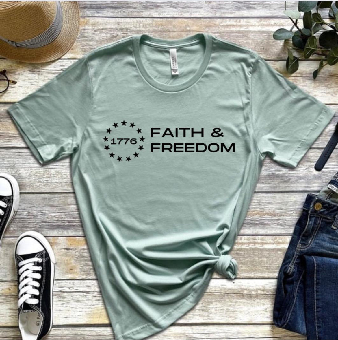 Dusty green crewneck t-shirt that says, “Faith and freedom” with a Betsy Ross flag star symbol and in the middle it says, “1776”.