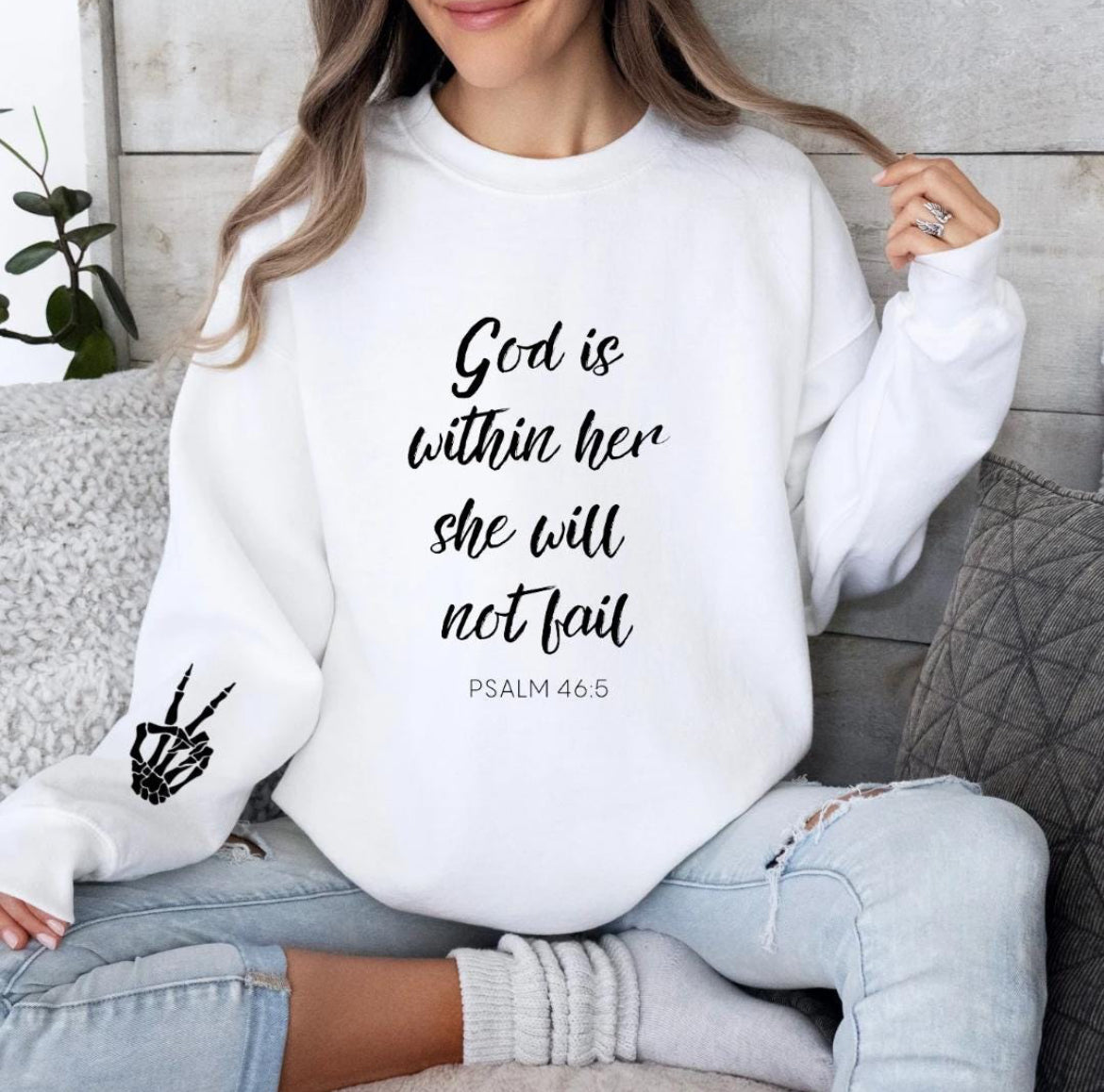 White crewneck sweatshirt that says, "God is within her she will not fail." Underneath in smaller letters it says, "Psalm 46 5." On the sleeve there is a bones peace sing.