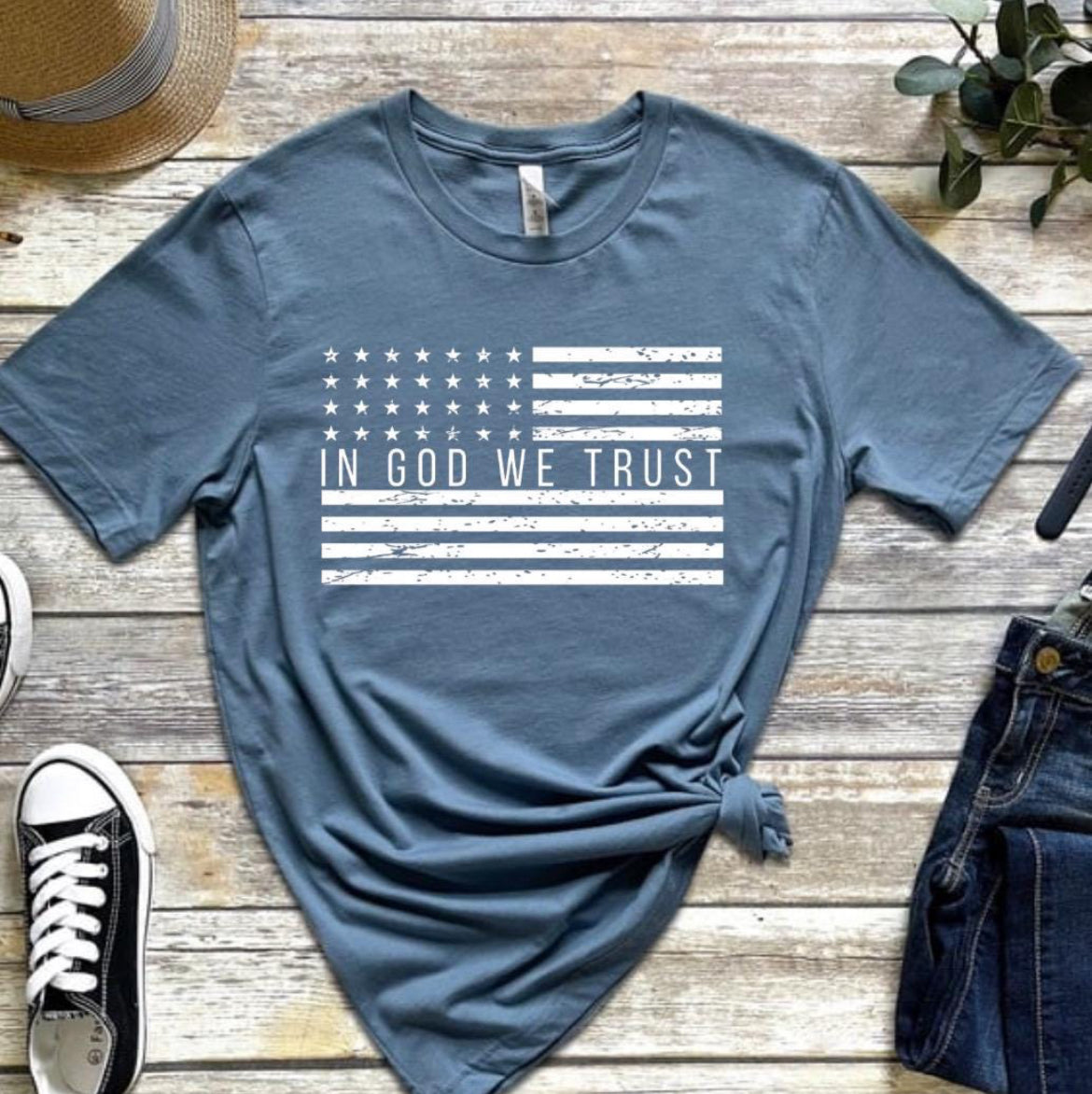 Steel blue crewneck t-shirt that has a distressed American flag. In the middle of the flag it says, “In God we trust.”