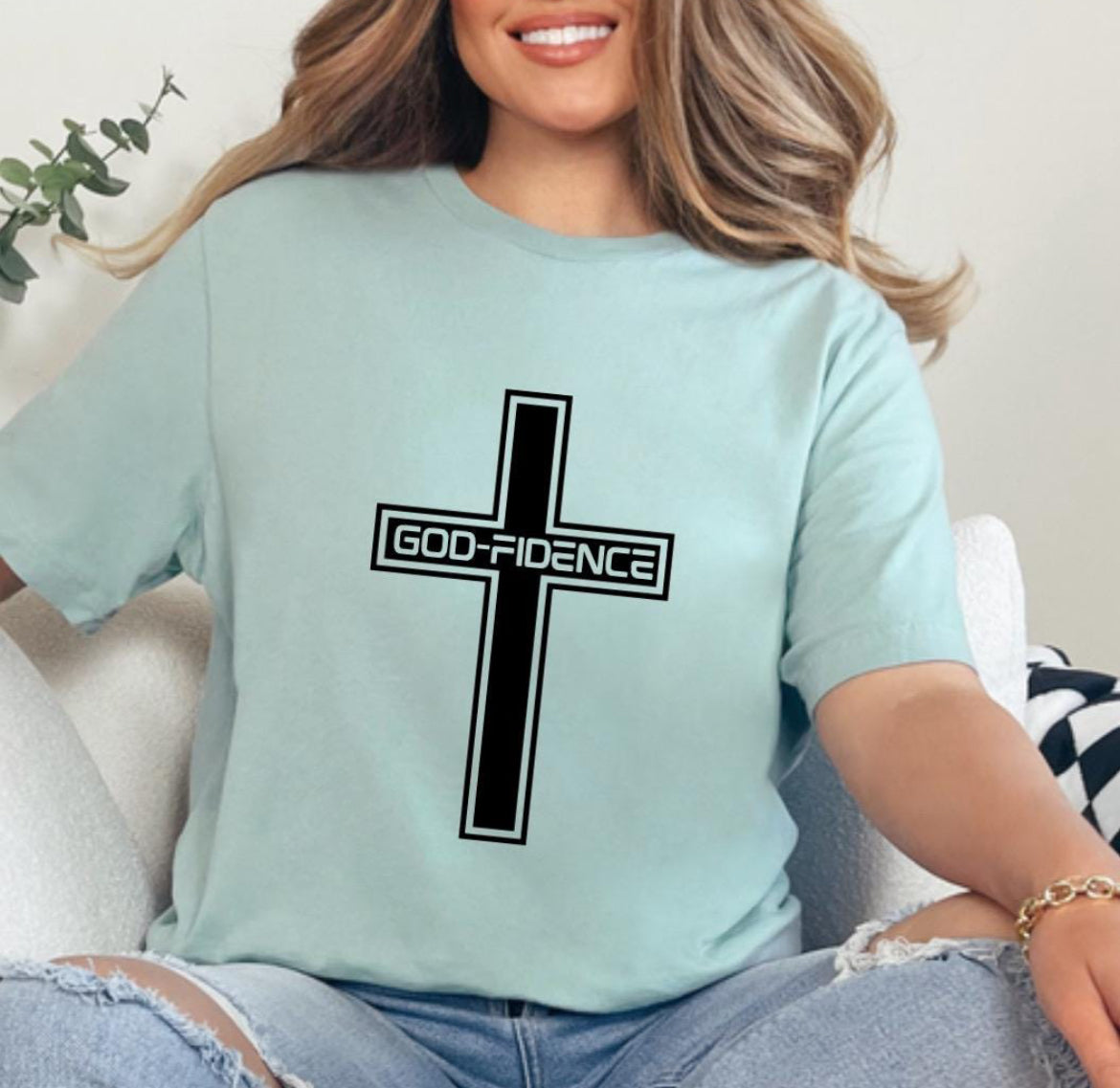 Dusty green crewneck t-shirt that says, “Godfidence” inside of a cross. 