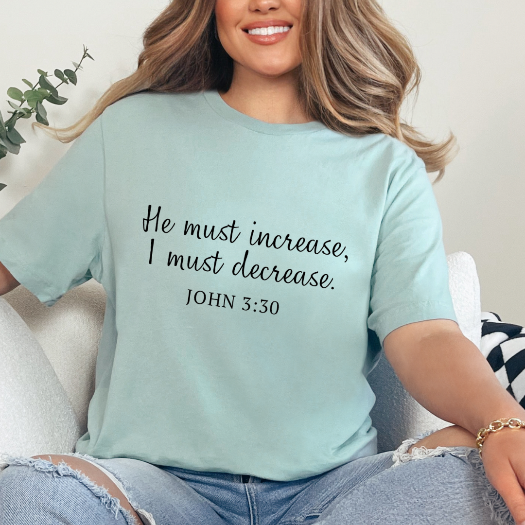 Dusty green crewneck t-shirt that says, “He must increase, I must decrease.” Underneath in smaller letters it says, “John 3 30.”