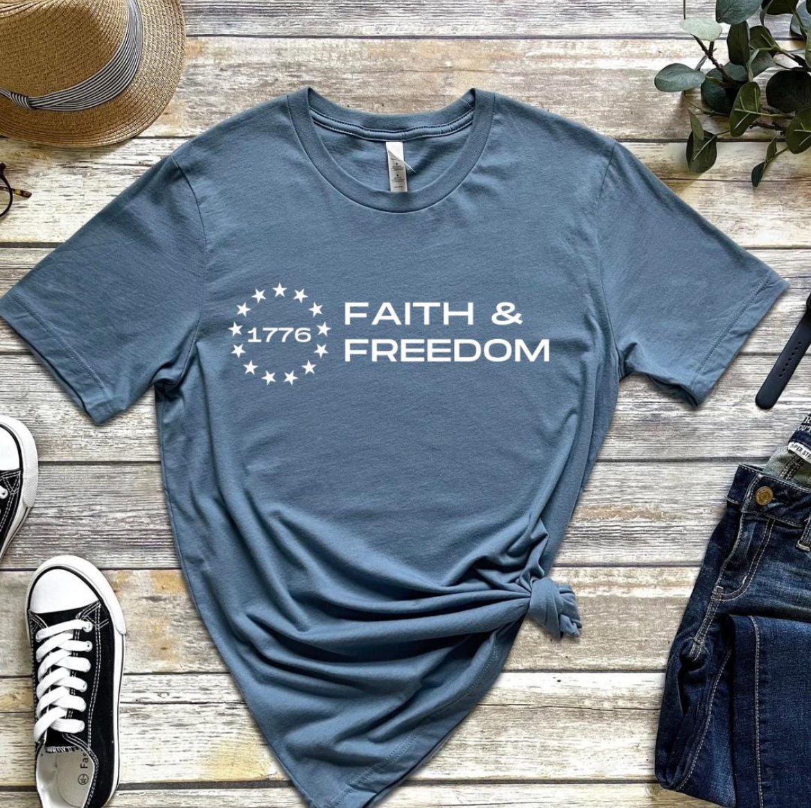 Steel blue crewneck t-shirt that says, “Faith and freedom” with a Betsy Ross flag star symbol and in the middle it says, “1776”.