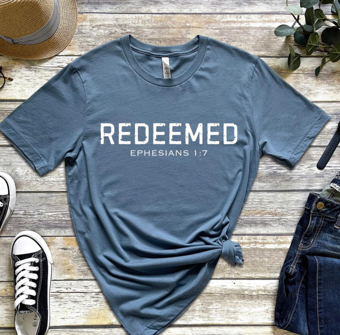 Steel blue Crewneck t-shirt that says, “redeemed” in large distressed letters. Underneath in smaller letters it says, “Ephesians 1 7.”