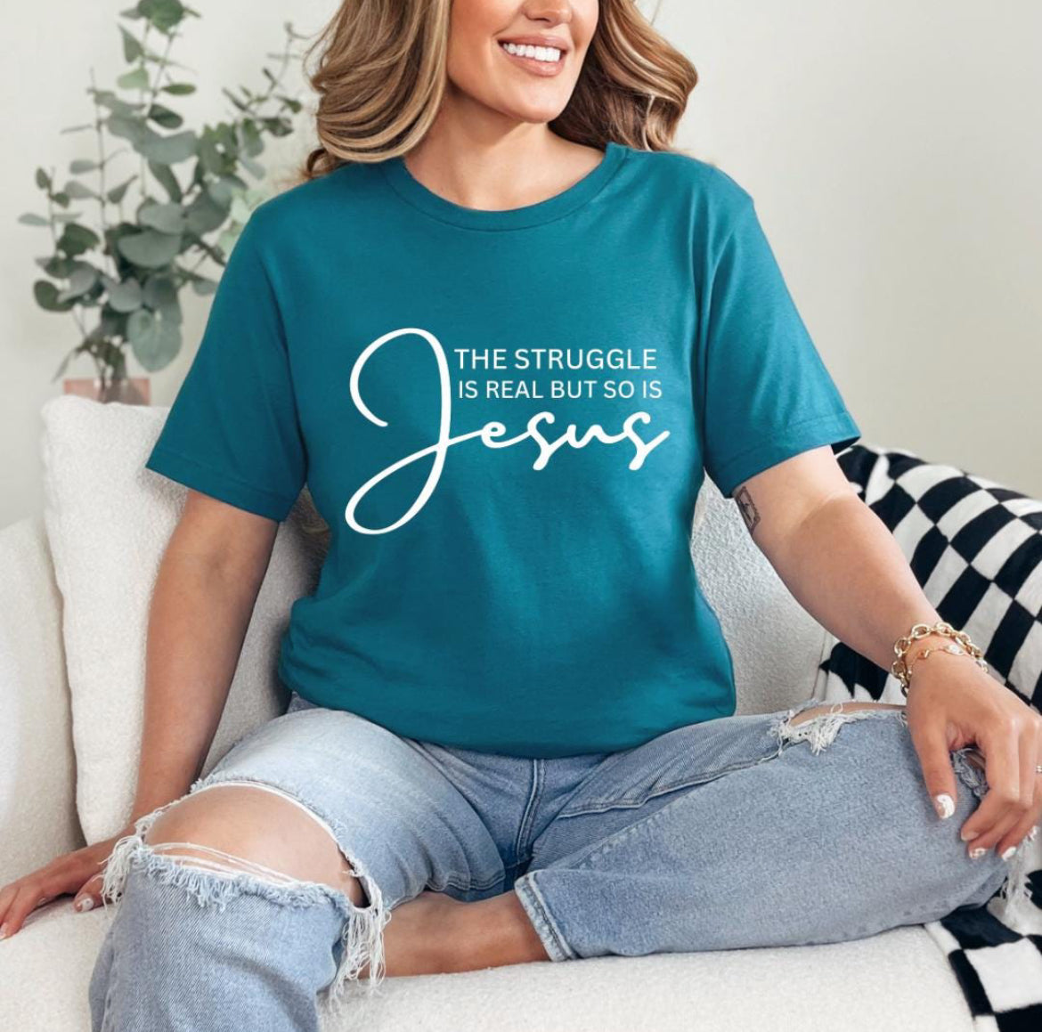 Deep teal crewneck t-shirt that says, “the struggle is real but so is Jesus”.