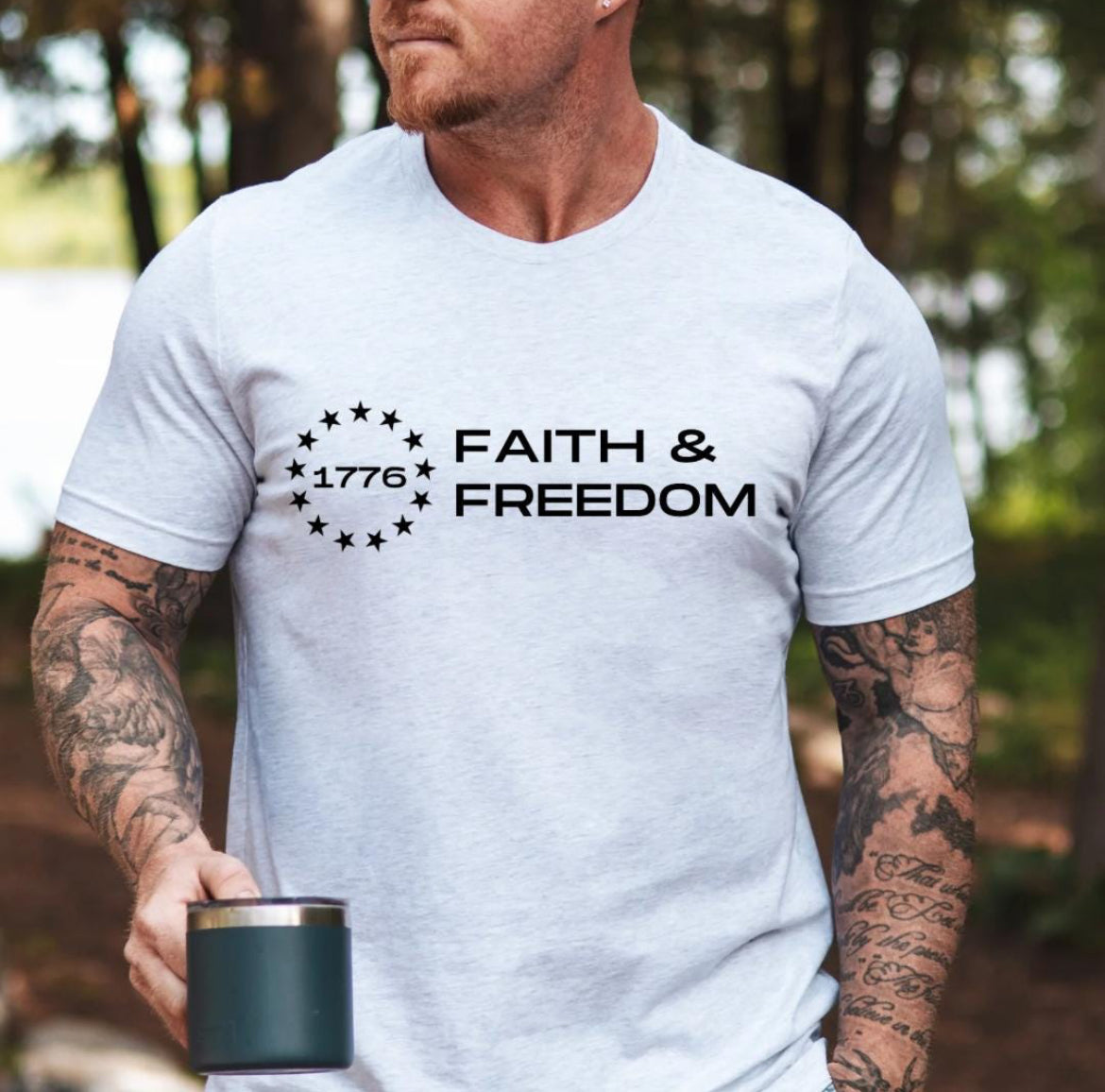 Ash white crewneck t-shirt that says, “Faith and freedom” with a Betsy Ross flag star symbol and in the middle it says, “1776”.