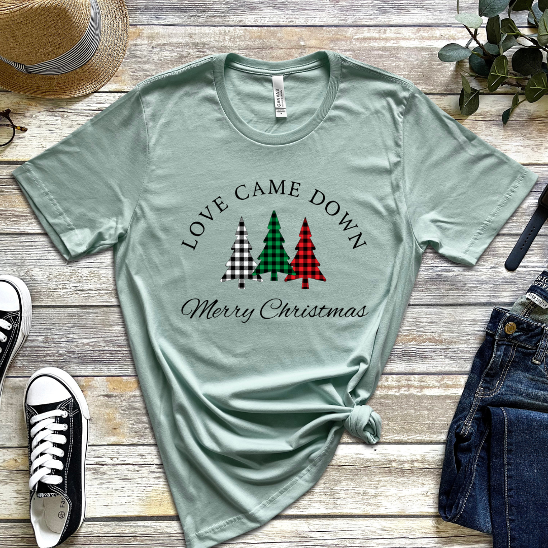 Dusty green crewneck t-shirt that says, "Love came down. Merry Christmas." It features 3 Christmas trees in buffalo plaid green, red and white.