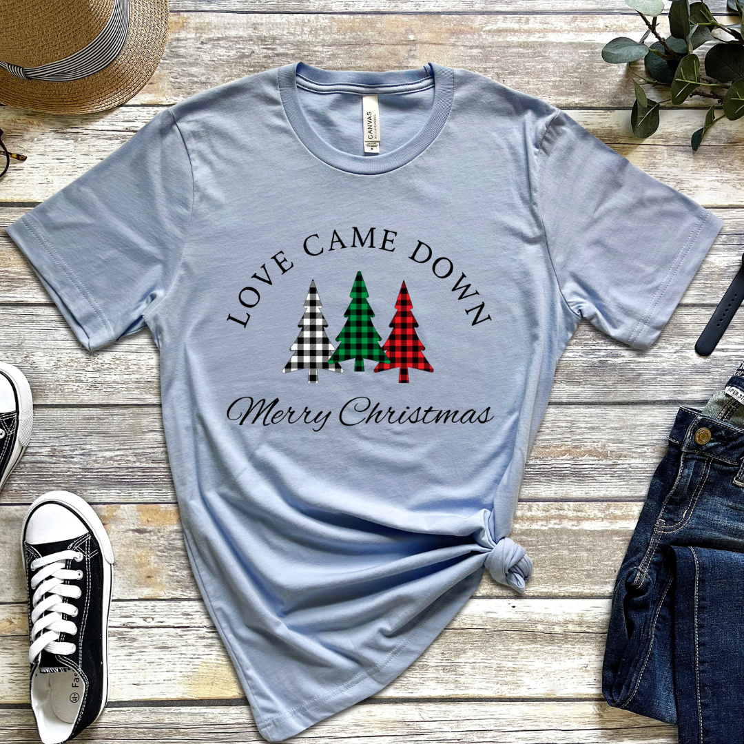 Baby blue crewneck t-shirt that says, "Love came down. Merry Christmas." It features 3 Christmas trees in buffalo plaid green, red and white.