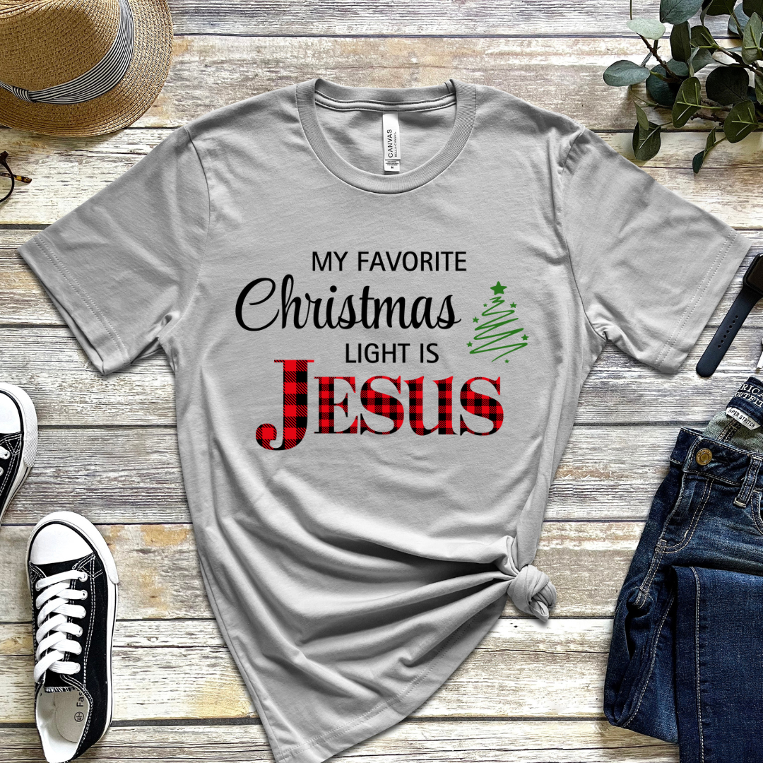 Grey crewneck t-shirt that says, "My favorite Christmas light is Jesus." Jesus is in red buffalo plaid, there is a Christmas tree graphic.