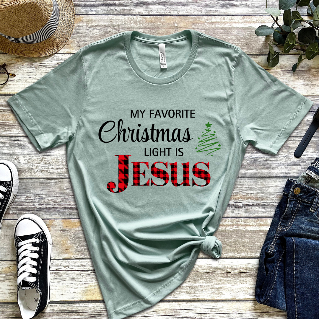 Dusty green crewneck t-shirt that says, "My favorite Christmas light is Jesus." Jesus is in red buffalo plaid, there is a Christmas tree graphic.