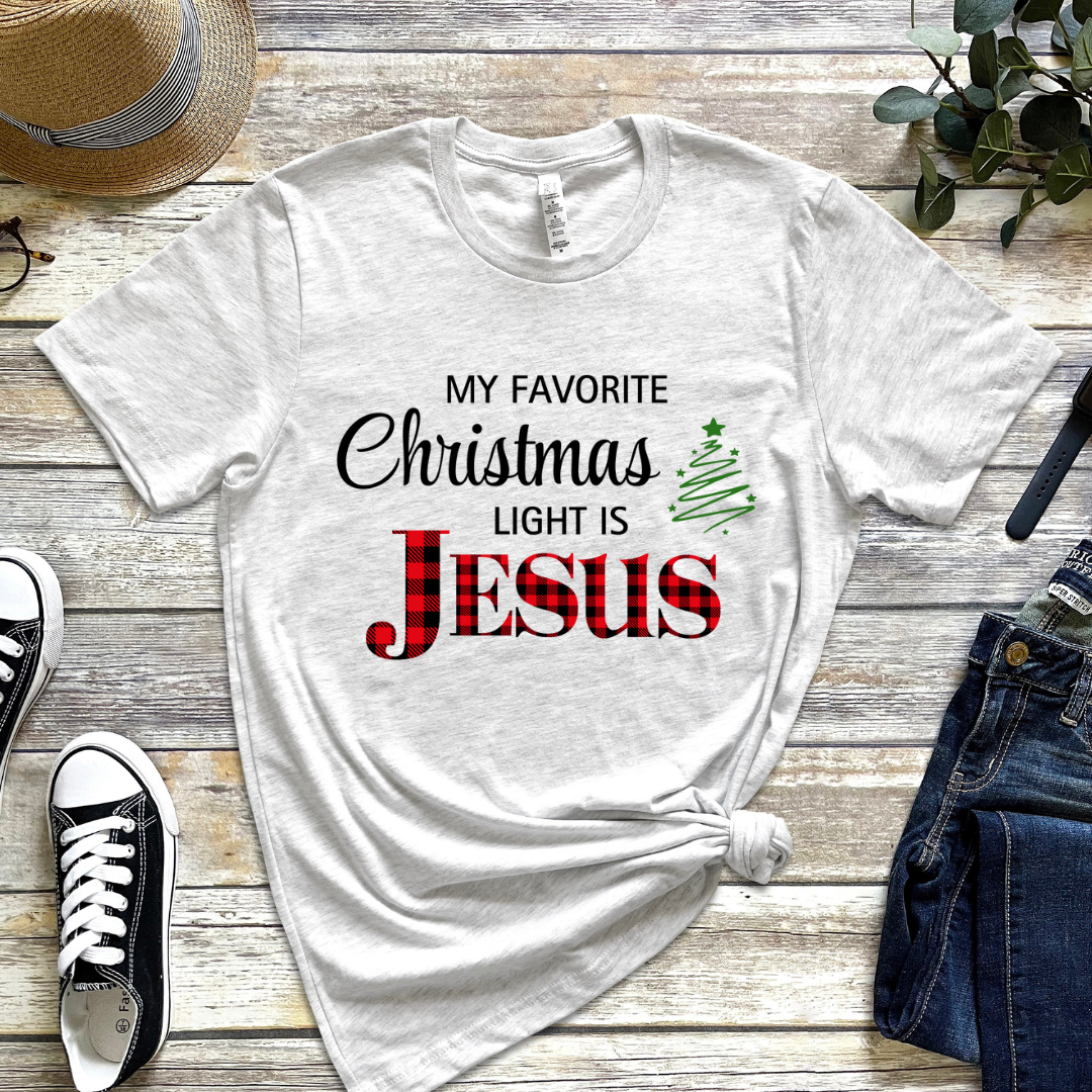 Ash white crewneck t-shirt that says, "My favorite Christmas light is Jesus." Jesus is in red buffalo plaid, there is a Christmas tree graphic.