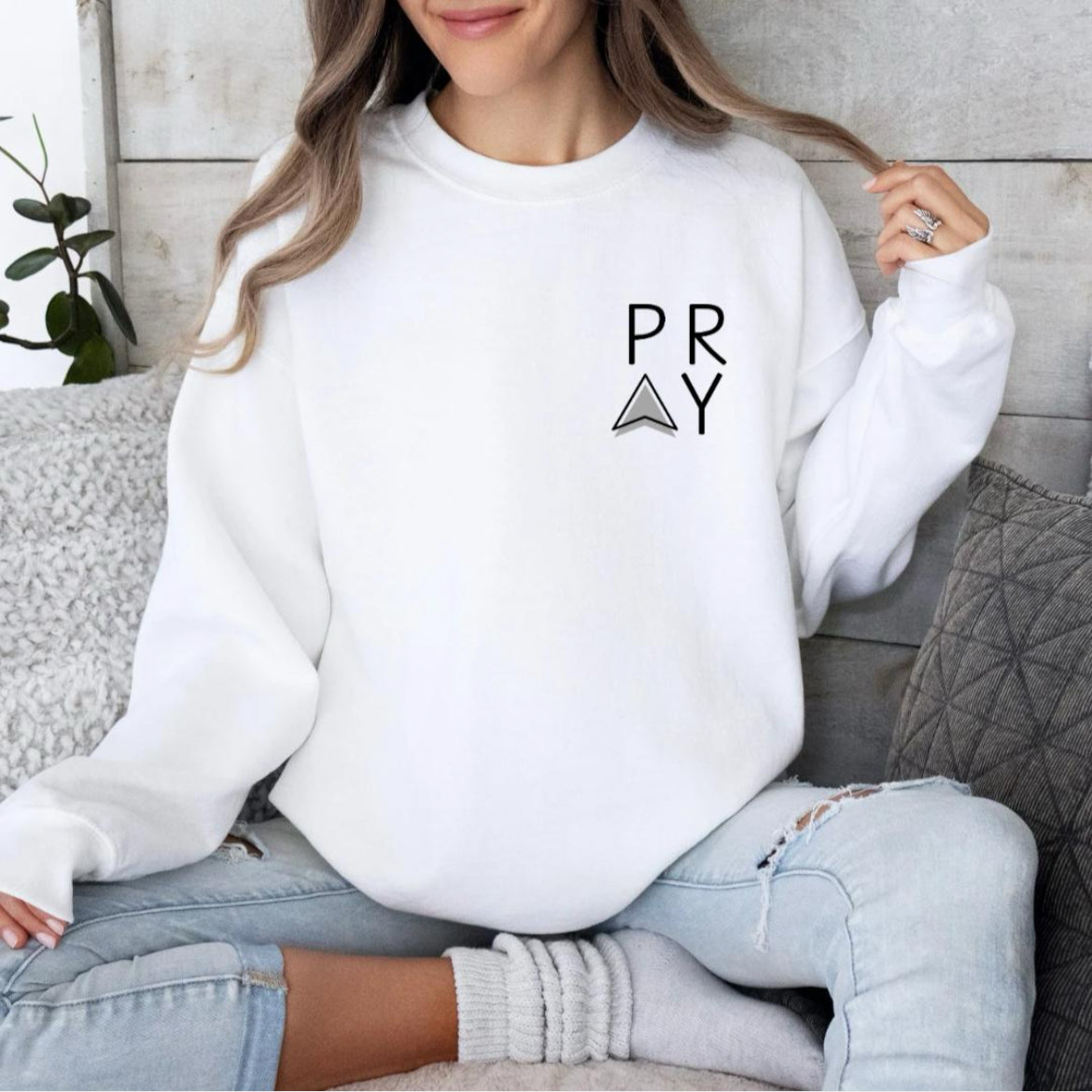 White crewneck sweatshirt that says, "PRAY" where the A is an arrow in the front left-hand corner of the hoodie.