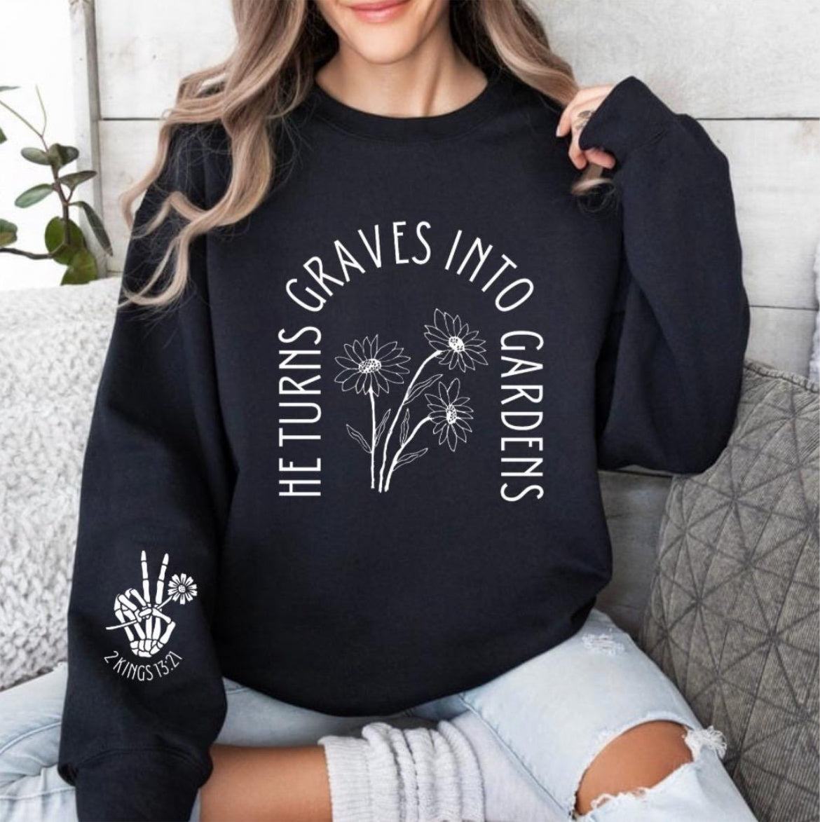 Black crewneck sweatshirt that says, "He turn graves into gardens." The words are in the shape of a grave. In the middle are flowers. On the sleeve is a peace sign bones hand holding a flower and underneath it says, "2 Kings 13 21."