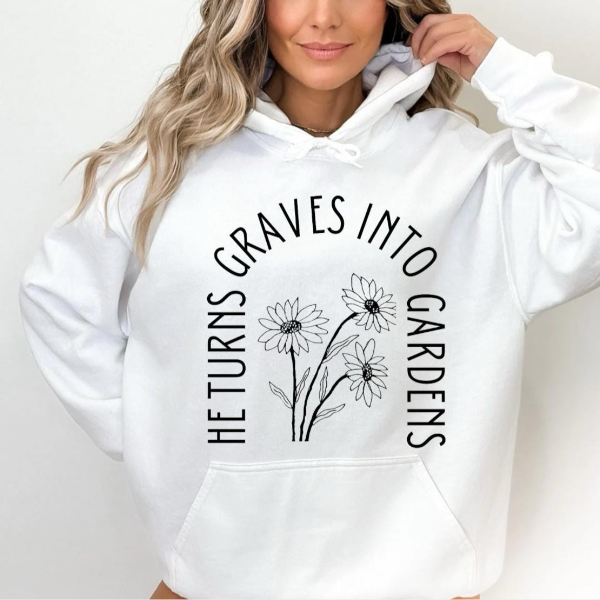 White hoodie that says, "He turn graves into gardens." The words are in the shape of a grave. In the middle are flowers. On the sleeve is a peace sign bones hand holding a flower and underneath it says, "2 Kings 13 21."