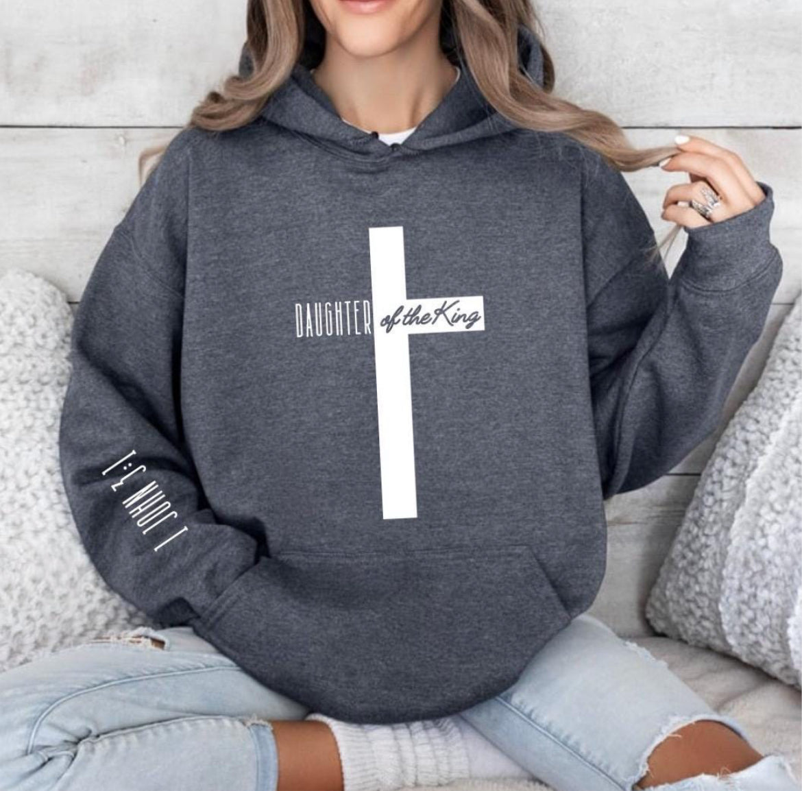 Dark grey heather hooded sweatshirt with a large cross that says, "Daughter of the King," On the sleeve it says, "1 John 3:1."