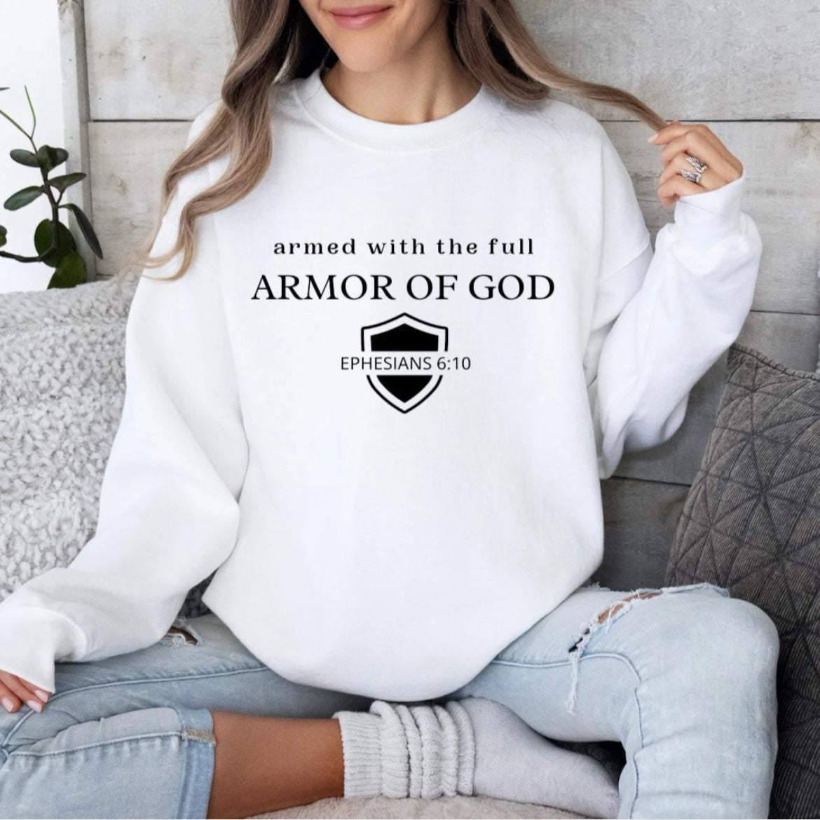 White crewneck sweatshirt that says, "armed with the full armor of God." Underneath is a shield and in the middle of the shield it says, "Ephesians 6 10."