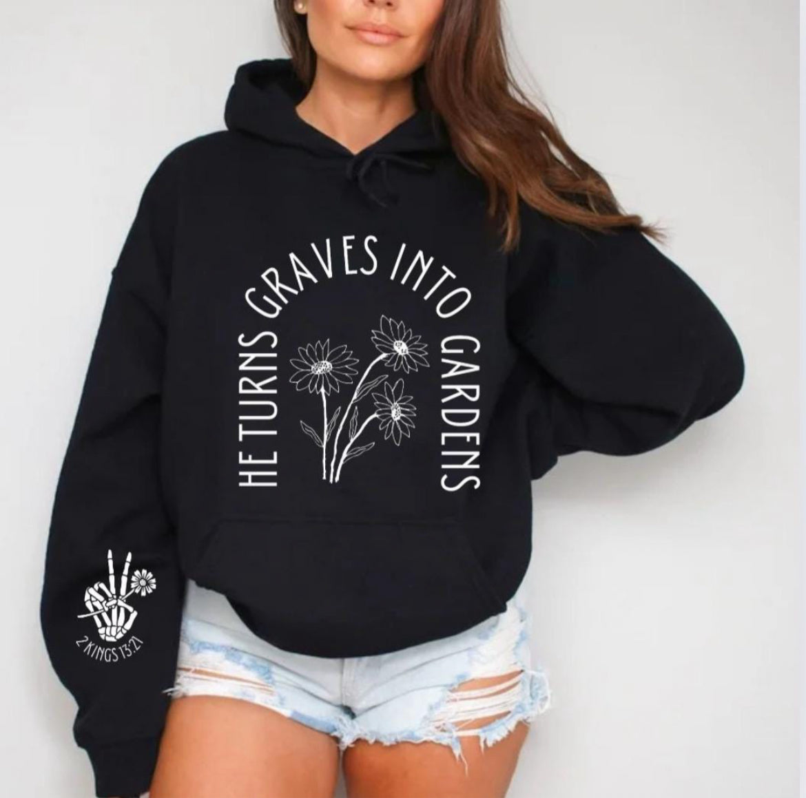 Black hoodie that says, "He turn graves into gardens." The words are in the shape of a grave. In the middle are flowers. On the sleeve is a peace sign bones hand holding a flower and underneath it says, "2 Kings 13 21."