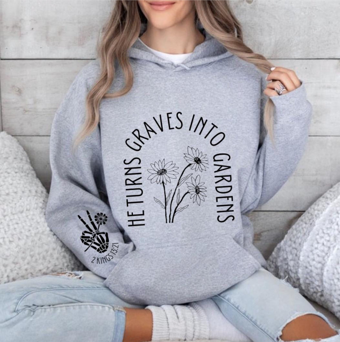 Light gray hoodie that says, "He turn graves into gardens." The words are in the shape of a grave. In the middle are flowers. On the sleeve is a peace sign bones hand holding a flower and underneath it says, "2 Kings 13 21."