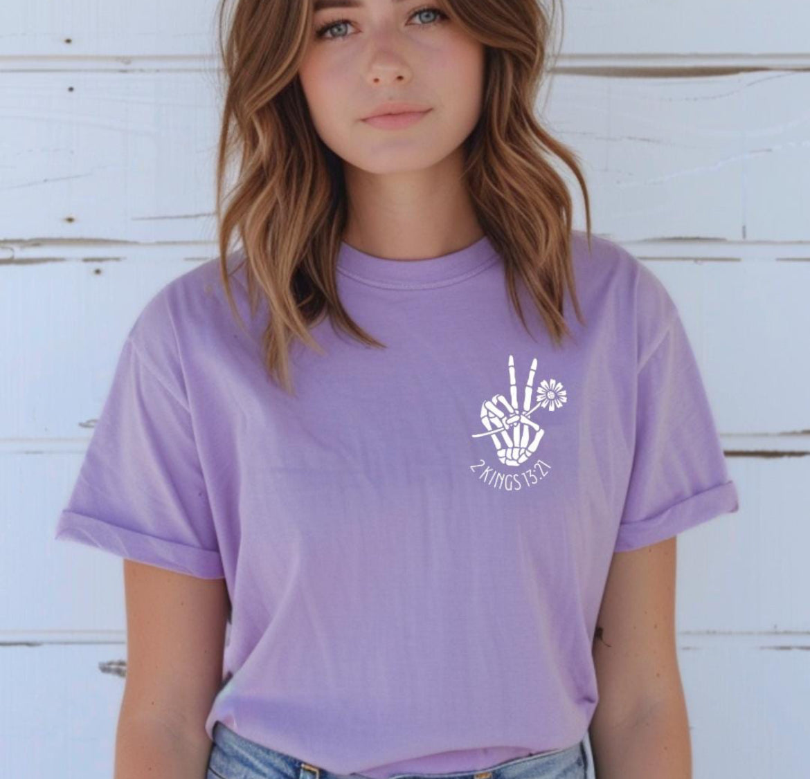 Lavender crewneck t-shirt that has a bones hand peace sign holding a flower. Underneath it says, “2 Kings 13 21.”